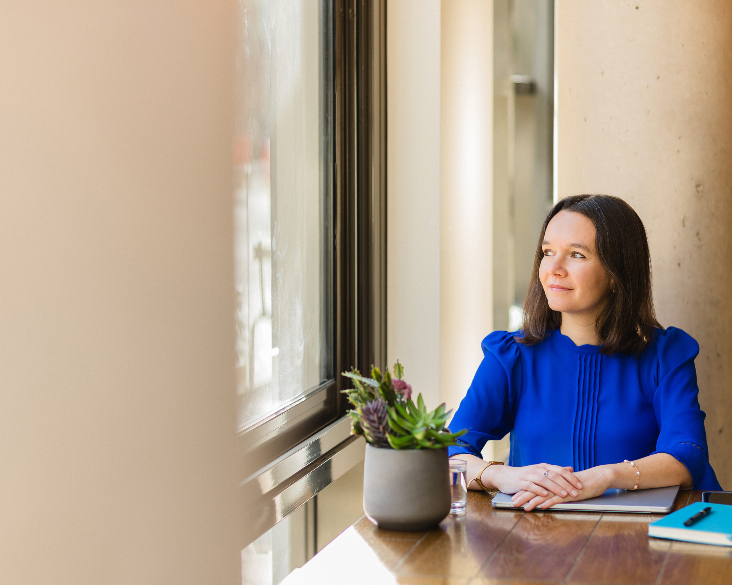 Laurence Galland looking out a window with a subtle smile, hands resting on a closed laptop